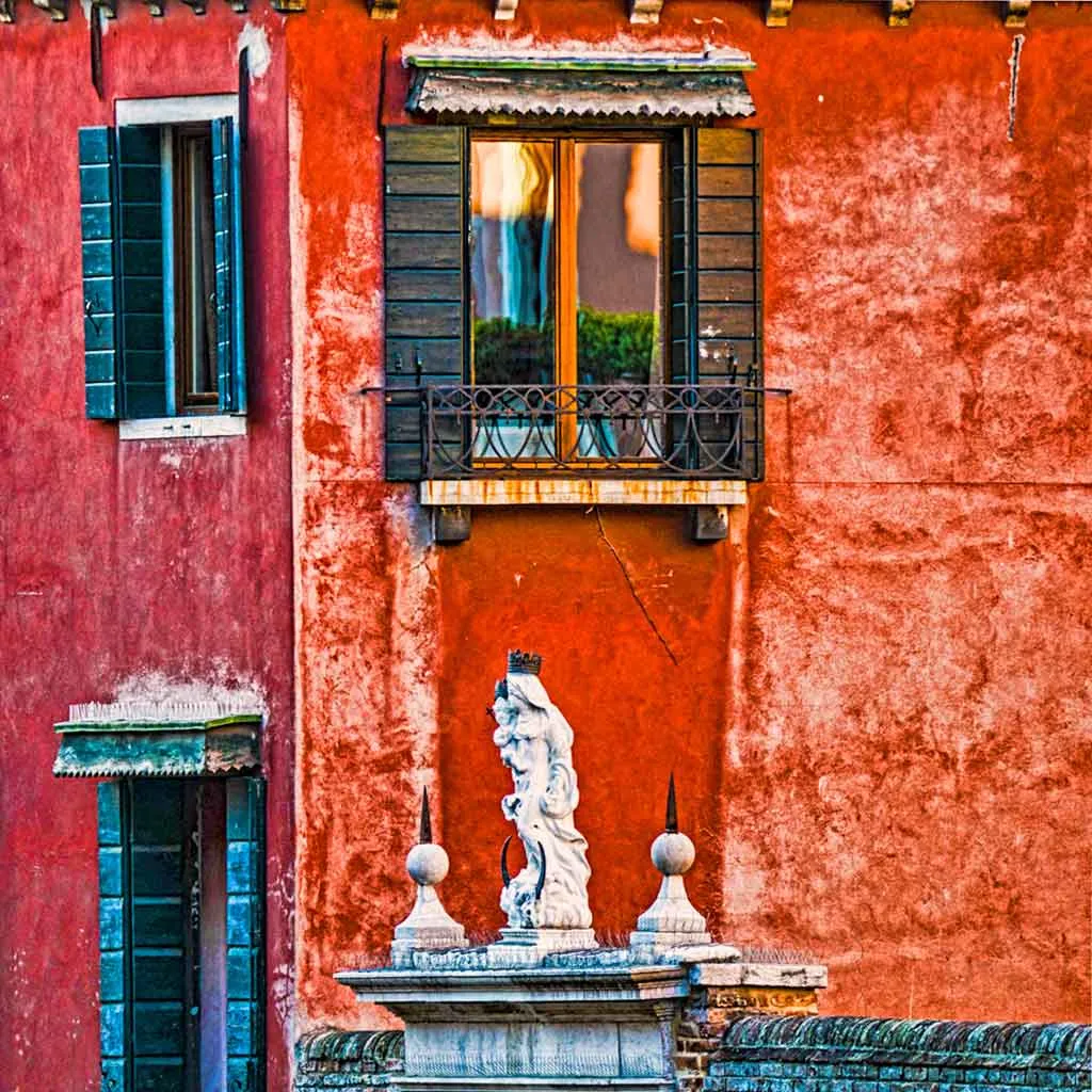 A decorative balcony with black shutters above a weathered red wall, featuring a white statue at the bottom center
