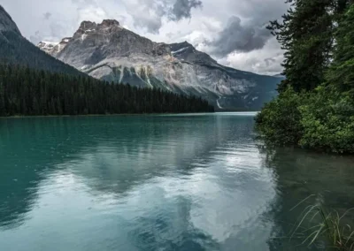 A serene lake reflects surrounding mountains and lush forests under a cloudy sky, creating a tranquil natural landscape