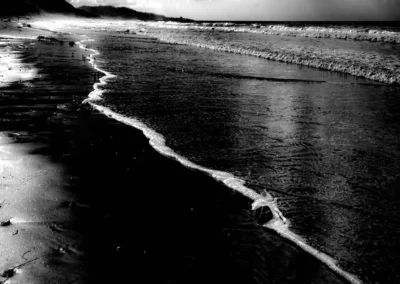 A dramatic black and white beach scene, featuring gentle waves lapping at the shore under moody, cloud-filled skies